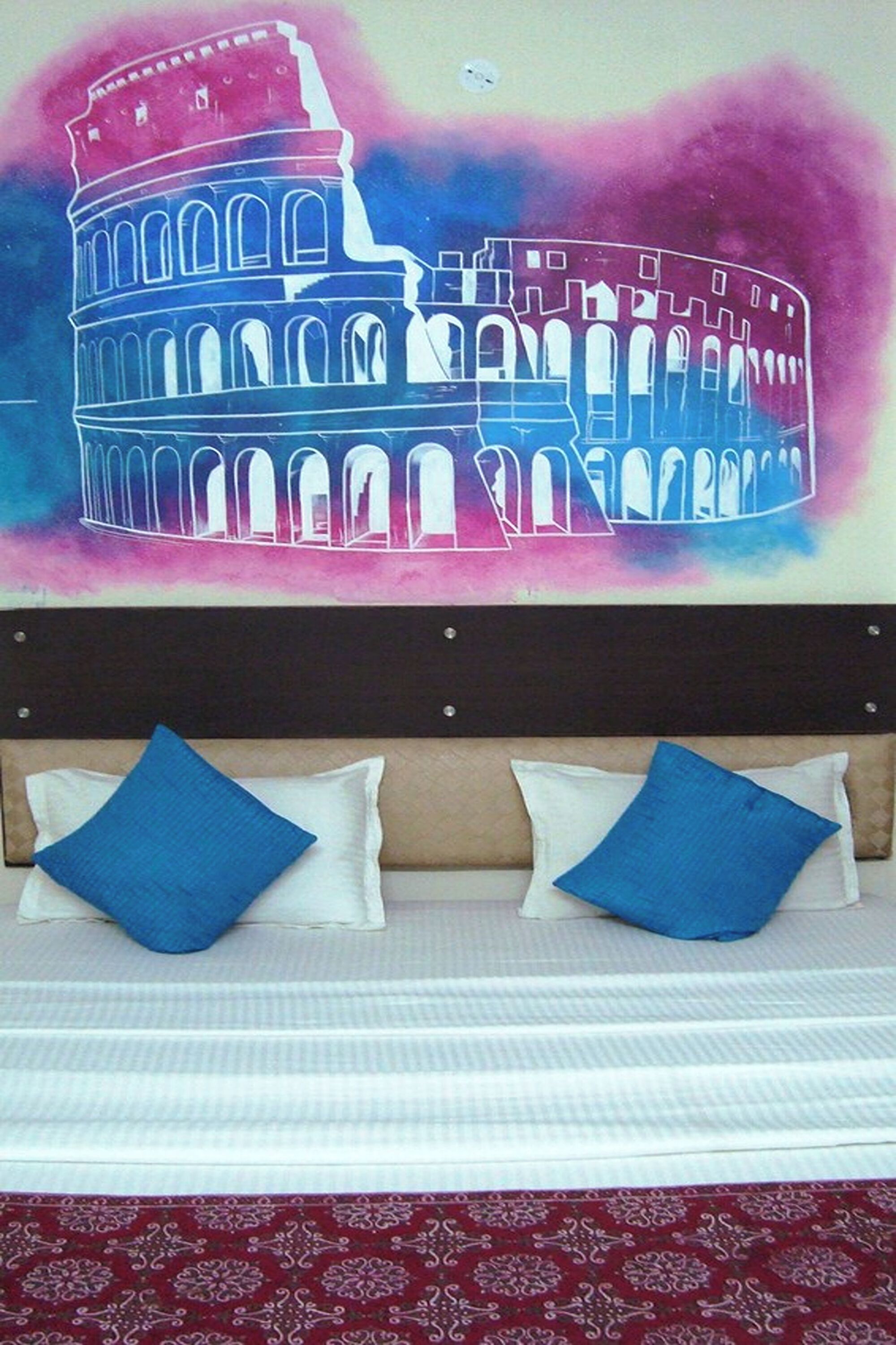 Uma cama com duas almofadas azuis e uma pintura colorida do Coliseu.