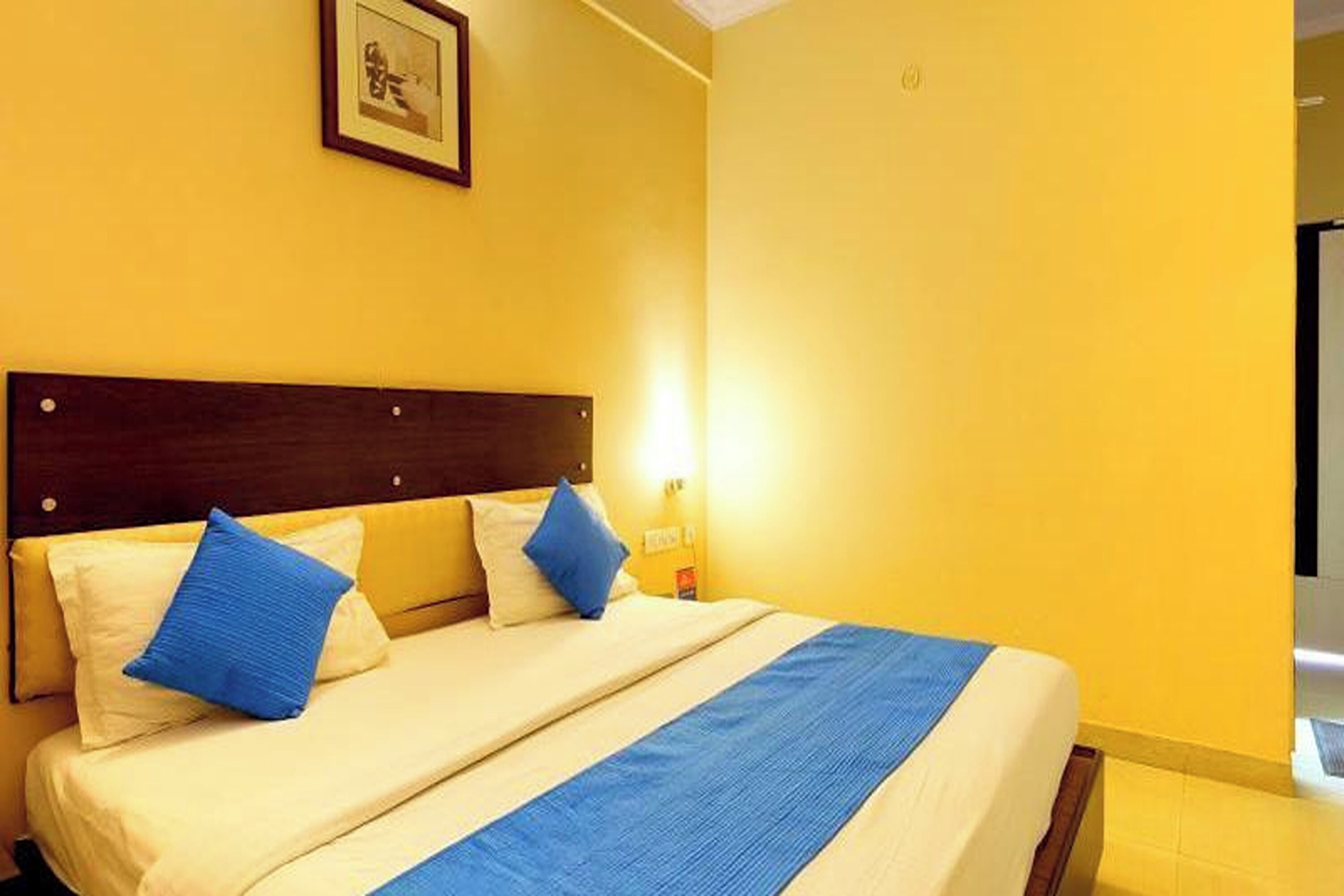 A hotel room with a bed, two pillows, a headboard, a wall-mounted picture, and a nightstand with a lamp.
