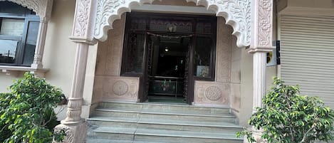 A building entrance with ornate carvings, a set of stairs leading up to it, and two potted plants on either side.