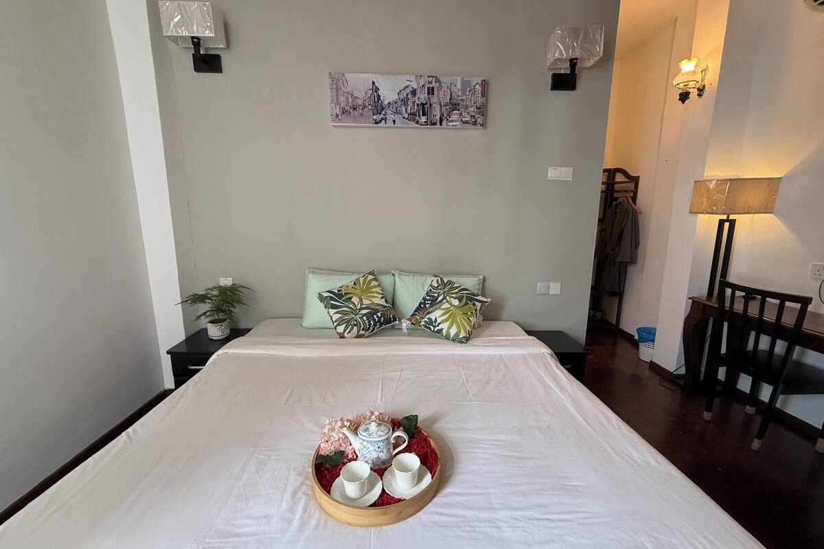 A neatly made bed with a tray of teaware and a painting above it.