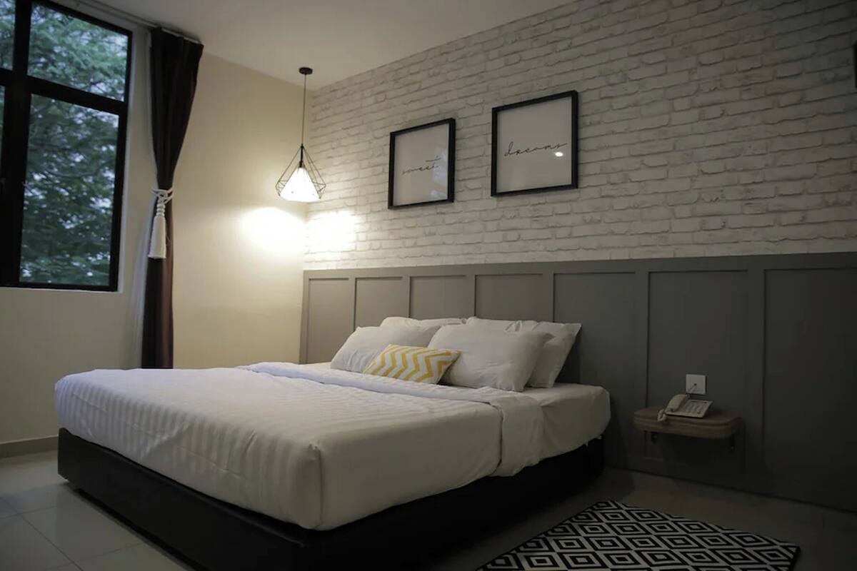 A modern bedroom with a white brick wall, a bed with white bedding, a hanging light fixture, and a small bedside table with a telephone.