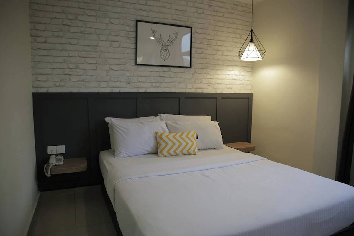 A neatly made bed with white bedding and a single decorative pillow, a small bedside table with a lamp, and a framed artwork on a brick-patterned wall.