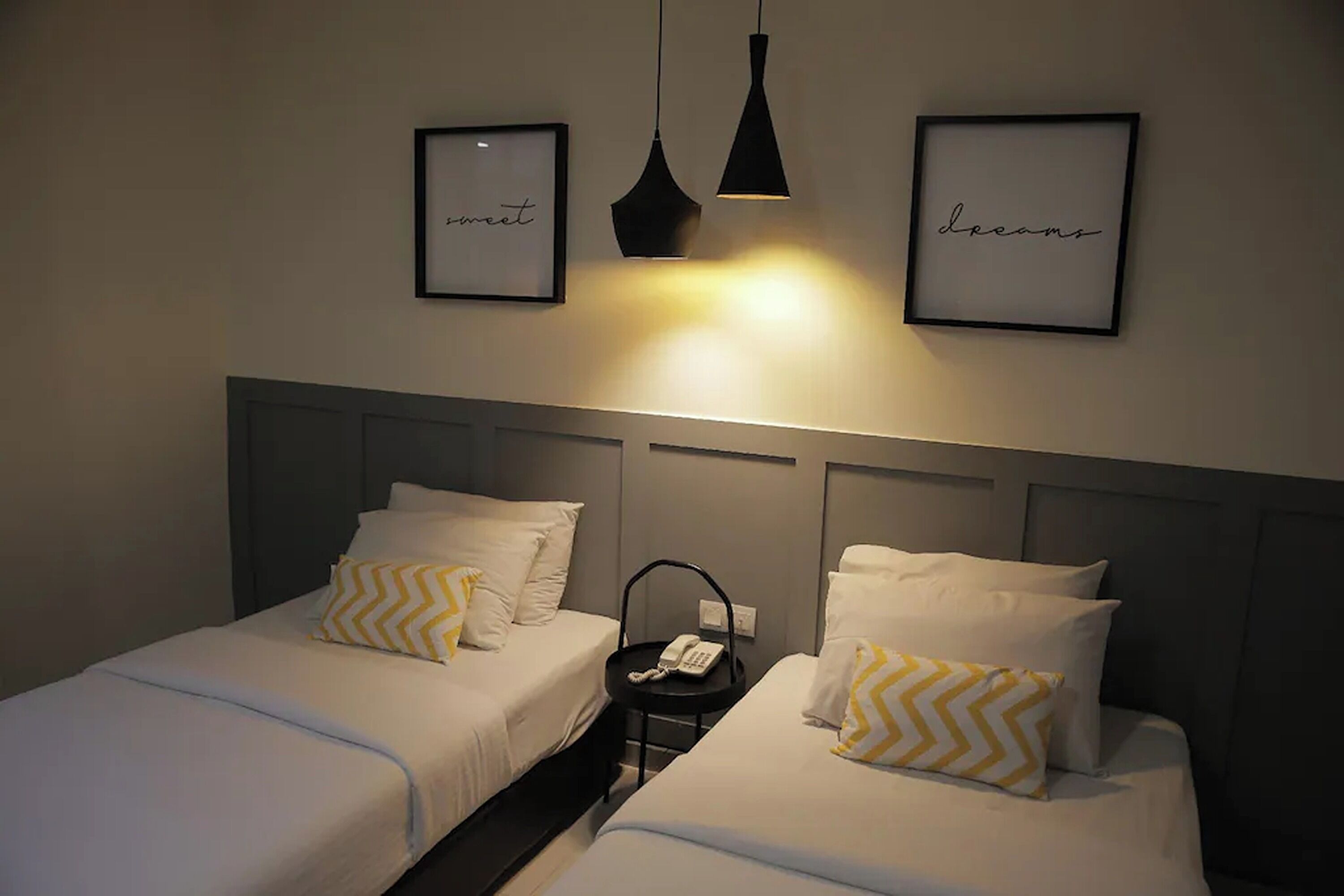 A hotel room with two single beds, each with white bedding and a yellow zigzag pillow. There are two framed pictures on the wall, one with the word "sweet" and another with "dreams." A black telephone is placed on a small table between the beds.