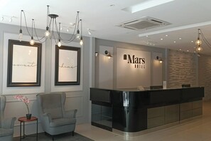 A modern hotel reception area with a black reception desk, framed artwork on the walls, and a seating area with a small table and chair. - Mars Hotel (Kuala Lumpur)