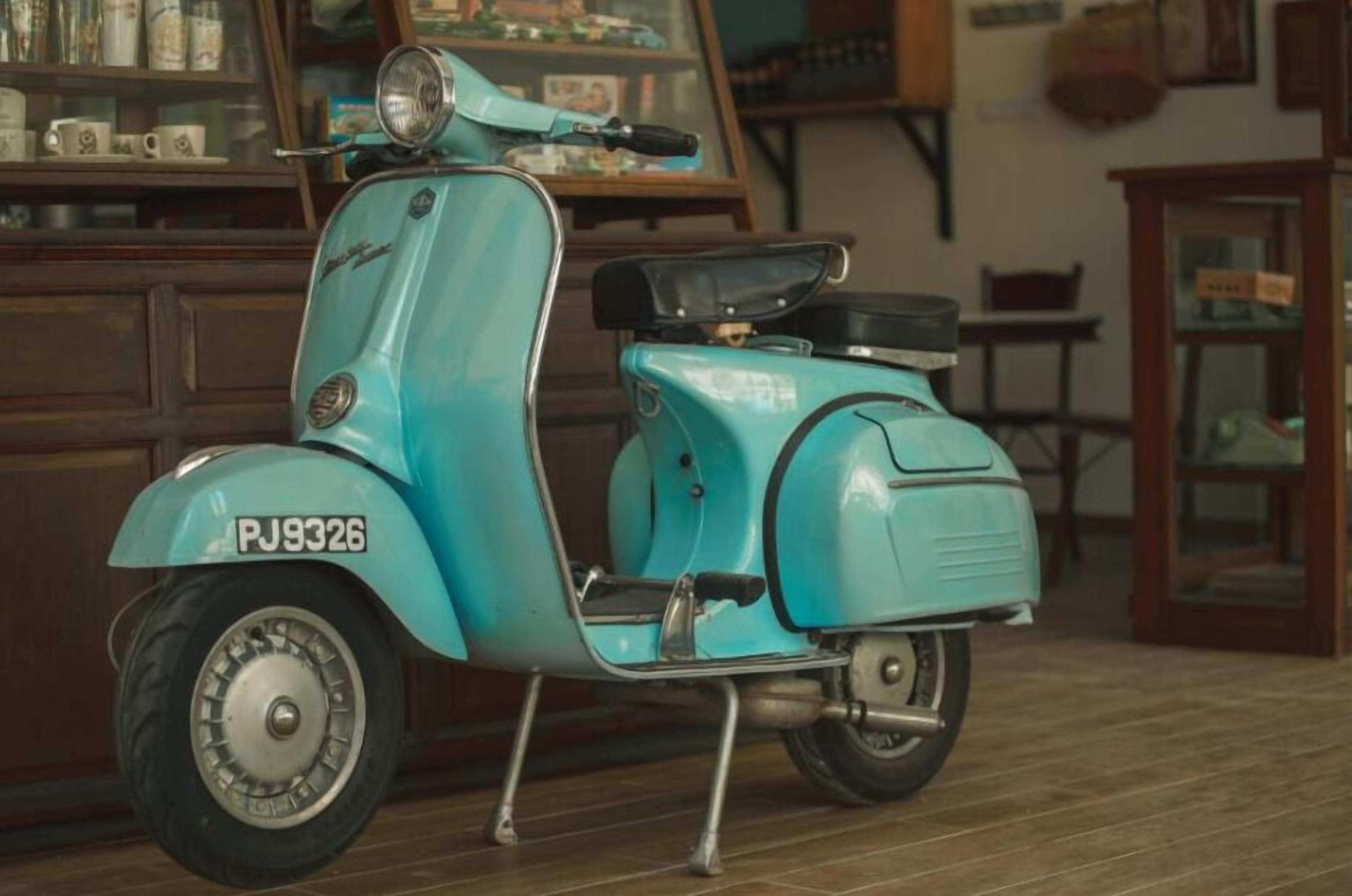 Un scooter Vespa vintage de couleur turquoise, portant le numéro d’immatriculation PJ9326, est garé à l’intérieur, avec des meubles en bois et un présentoir en verre en arrière-plan.