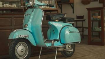 Un scooter Vespa vintage de couleur turquoise, portant le numéro d’immatriculation PJ9326, est garé à l’intérieur, avec des meubles en bois et un présentoir en verre en arrière-plan.