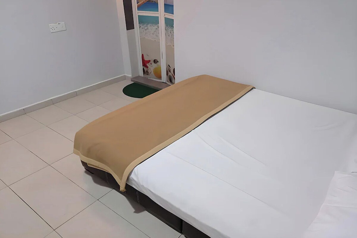 A bedroom with a bed, a small green mat, and a white wall with a power outlet.