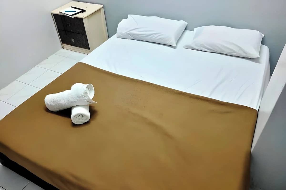 Hotellihuone, jossa on sänky, valkoiset tyynyt, ruskea peitto, valkoinen pyyhe ja pieni yöpöytä, jossa on musta laatikko.