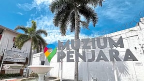A sign for 'Muzium Penjara' with a colorful location pin, a fountain, and a white wall with barbed wire.