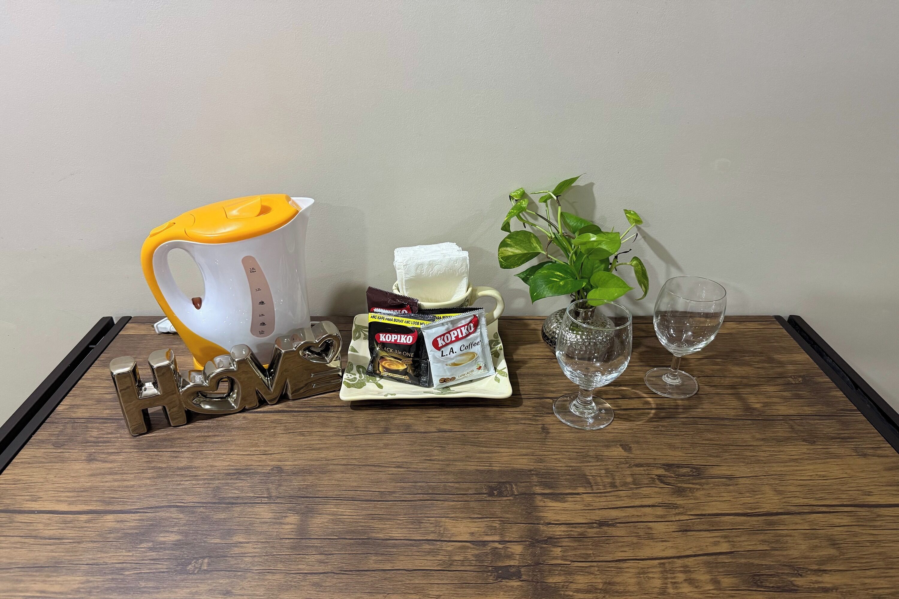 A wooden table with a coffee maker, a packet of Kopiko coffee, a small plant, and two wine glasses.