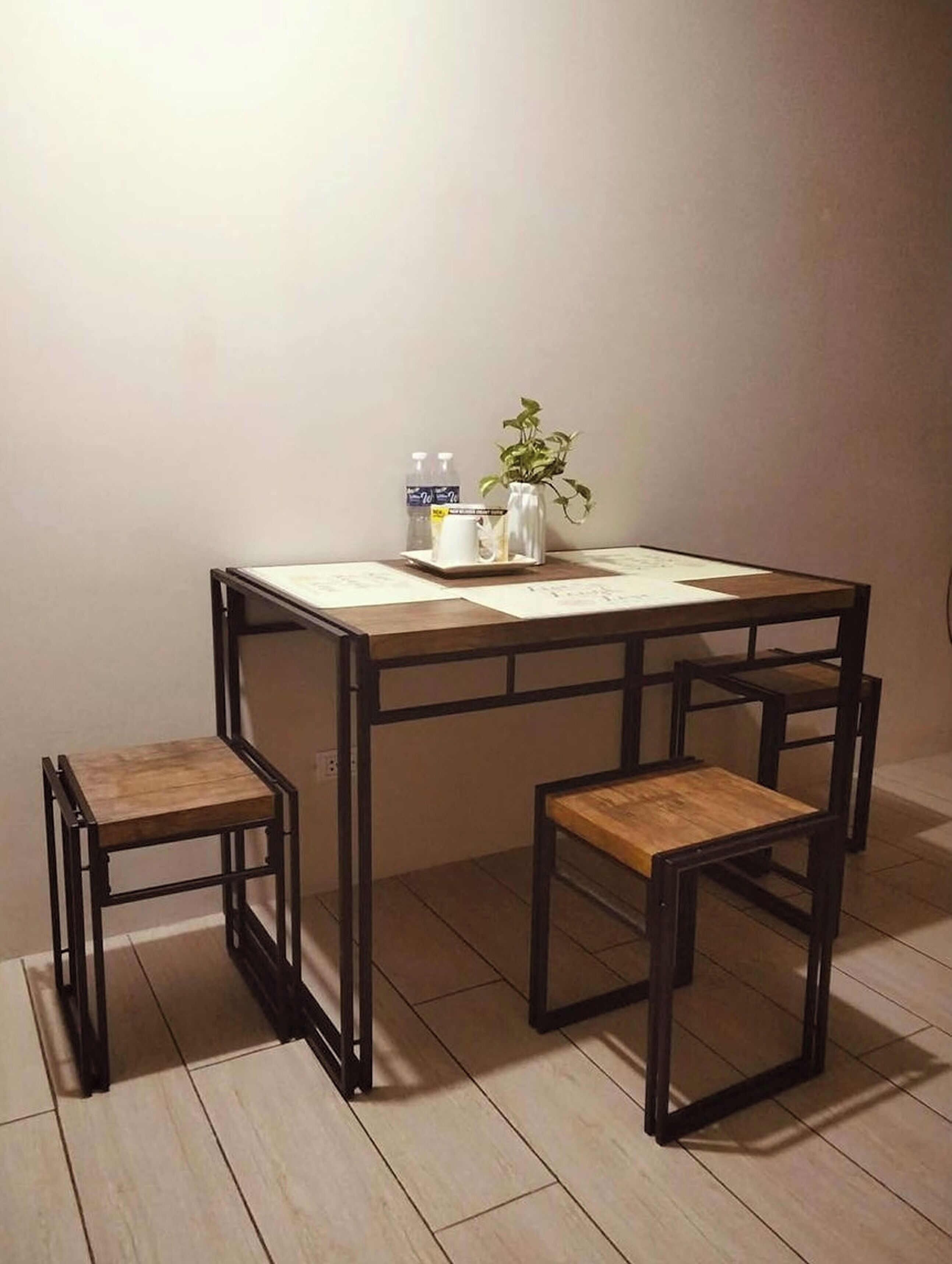 Una pequeña mesa cuadrada con tapa de madera y estructura de metal negro, acompañada por dos sillas a juego. Sobre la mesa se encuentra un florero blanco con una planta y una botella de agua.