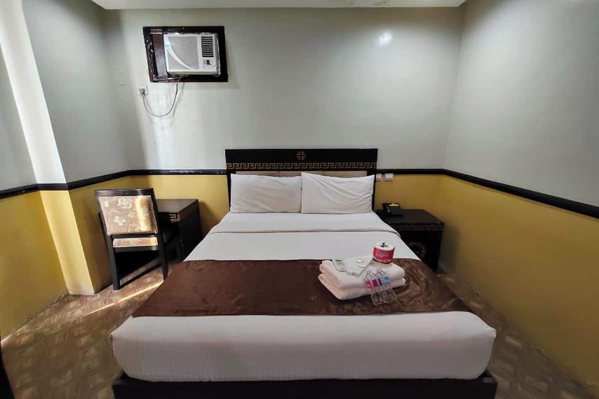 A hotel room with a bed, a nightstand, a chair, and an air conditioning unit on the wall.
