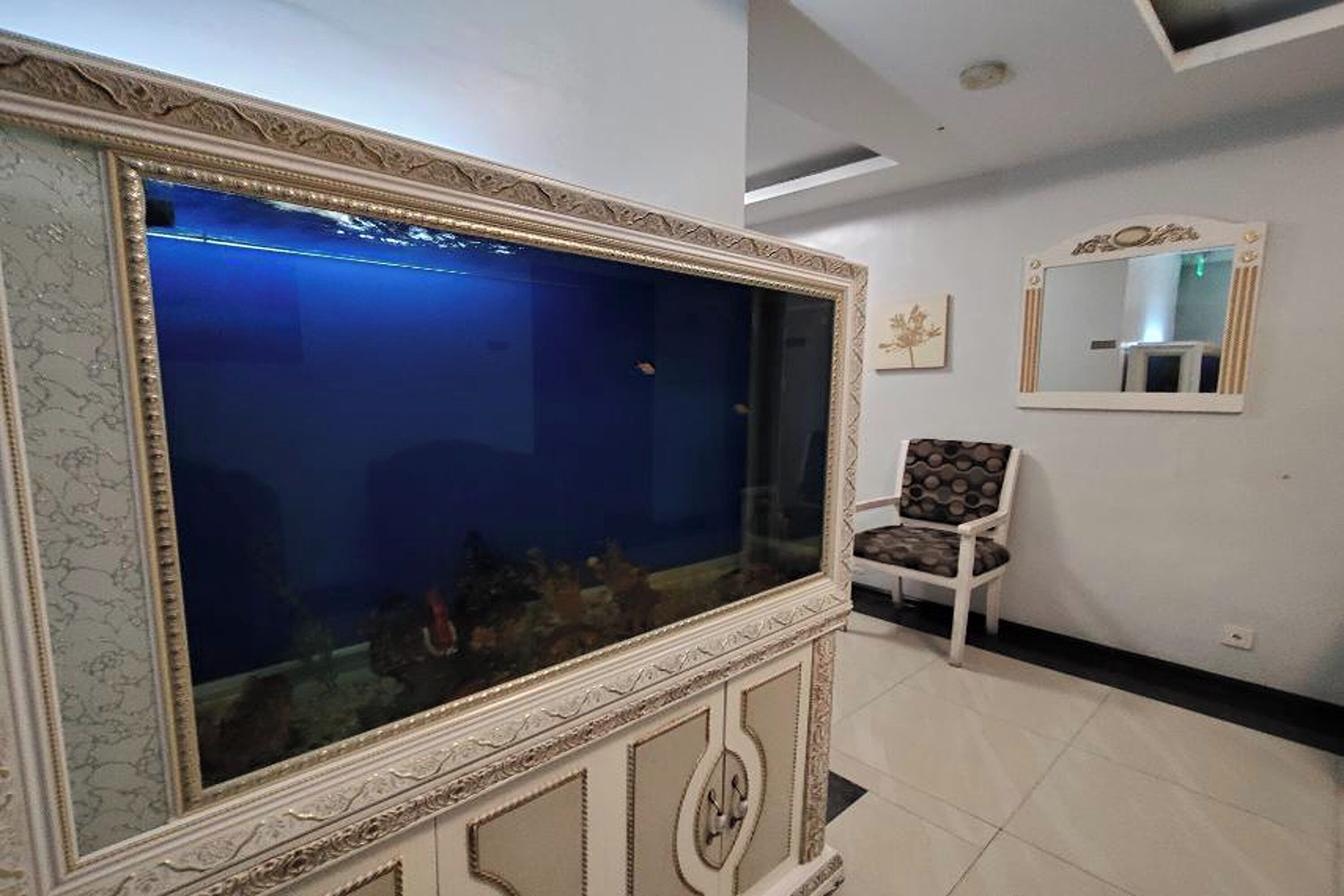 A room with a large ornate fish tank, a white chair, and a mirror on the wall.