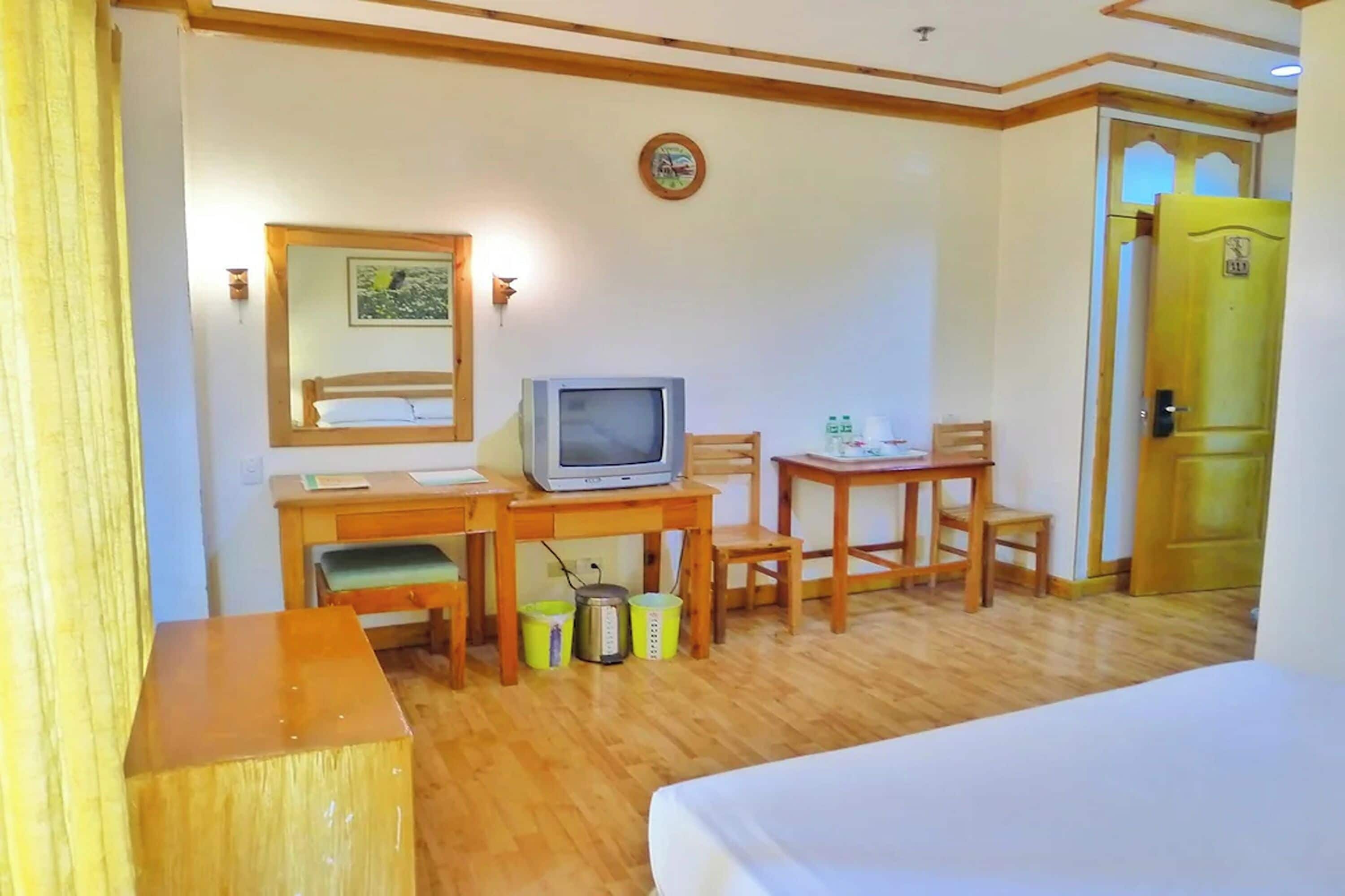 A hotel room with a bed, a desk with a television, a chair, a small table, and a wooden cabinet.