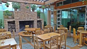 Ένα εστιατόριο με ξύλινα τραπέζια και καρέκλες, ένα τζάκι από πέτρα και φυτά σε γλάστρες.