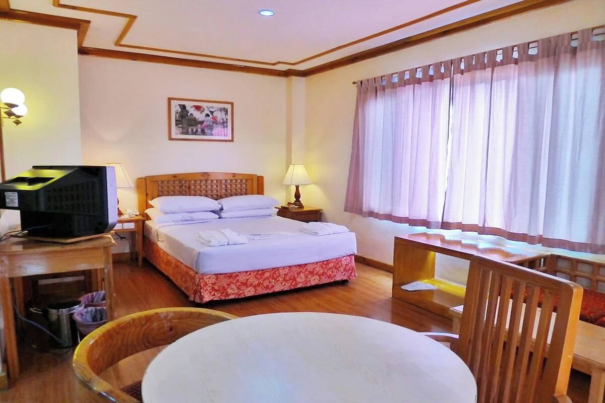 A hotel room with a bed, a desk with a lamp, a chair, a television, and a window with curtains.