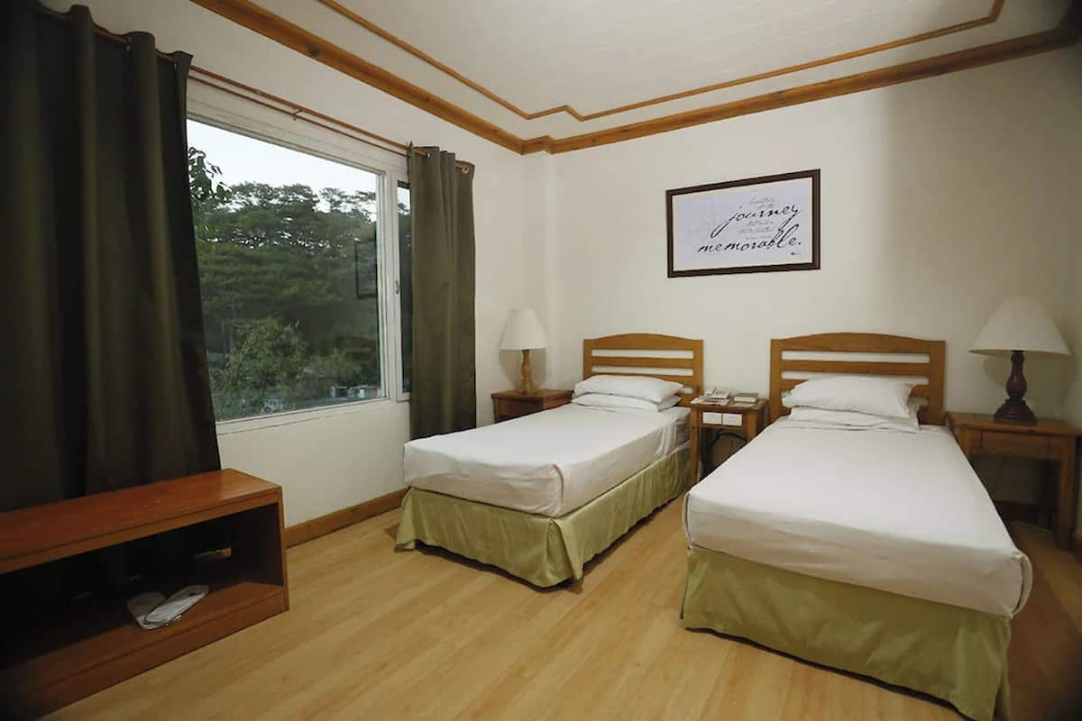 A room with two single beds, a wooden bench, a window with curtains, and a framed quote on the wall.