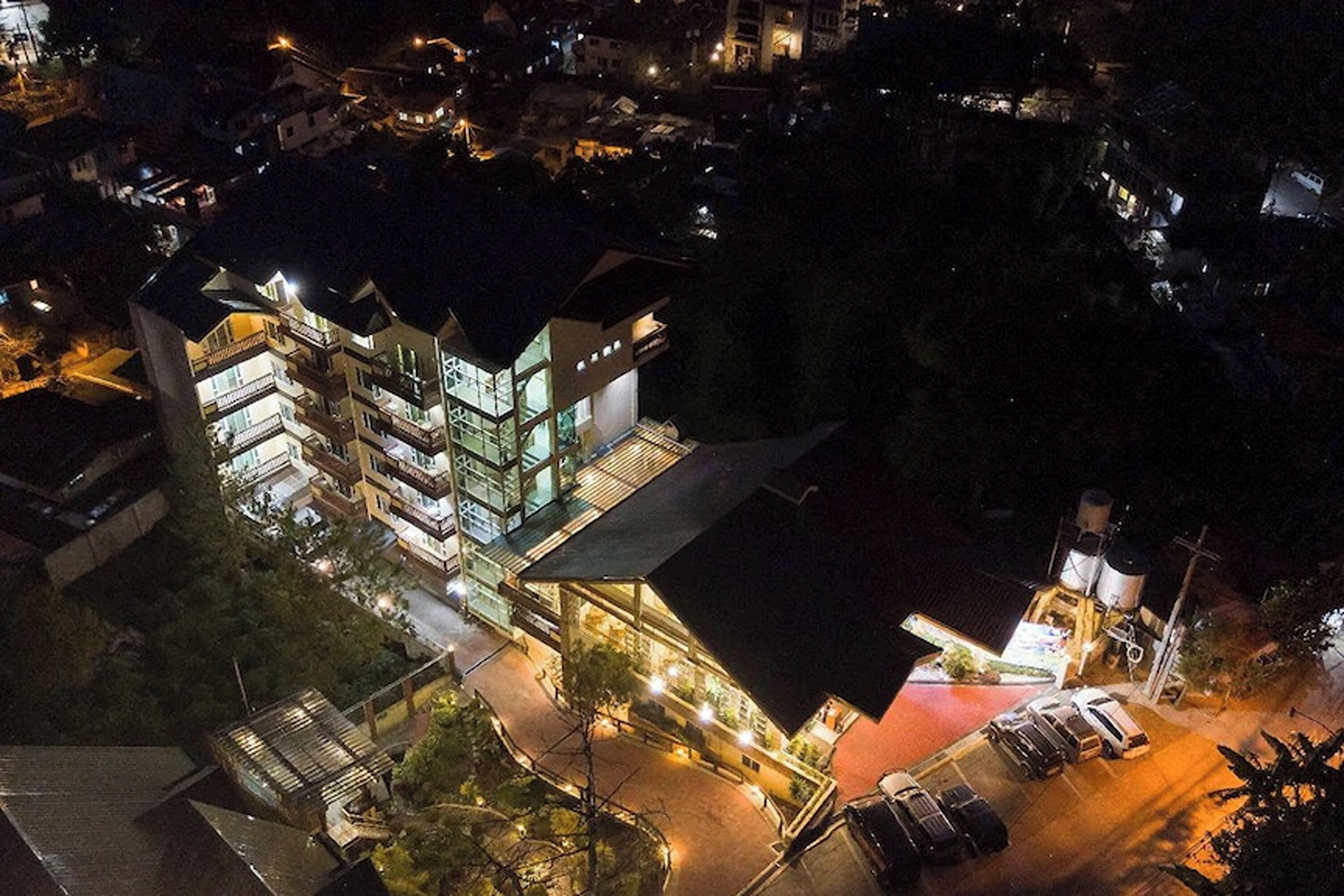 An aerial view of a modern hotel with multiple floors, a parking area, and surrounding buildings.