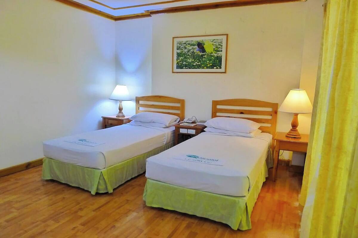 A hotel room with two single beds, wooden headboards, bedside tables with lamps, a framed picture on the wall, and a wooden floor.