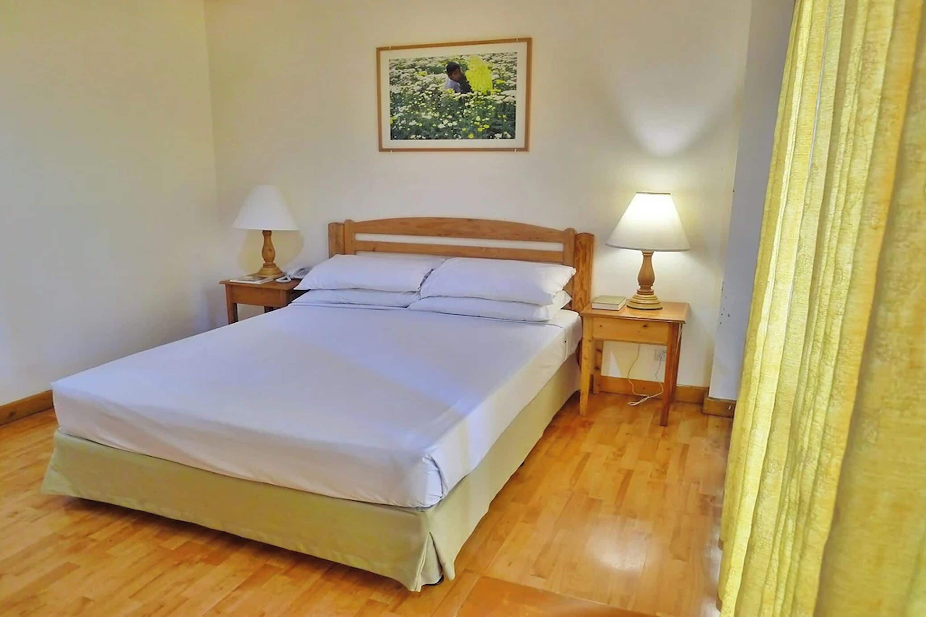 Una cama bien hecha en una habitación con suelo de madera, dos mesitas de noche con lámparas y un cuadro enmarcado en la pared.