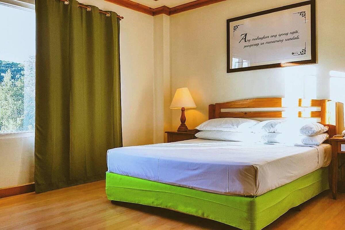A neatly made bed with a green bed skirt, a bedside lamp, and a framed quote on the wall.