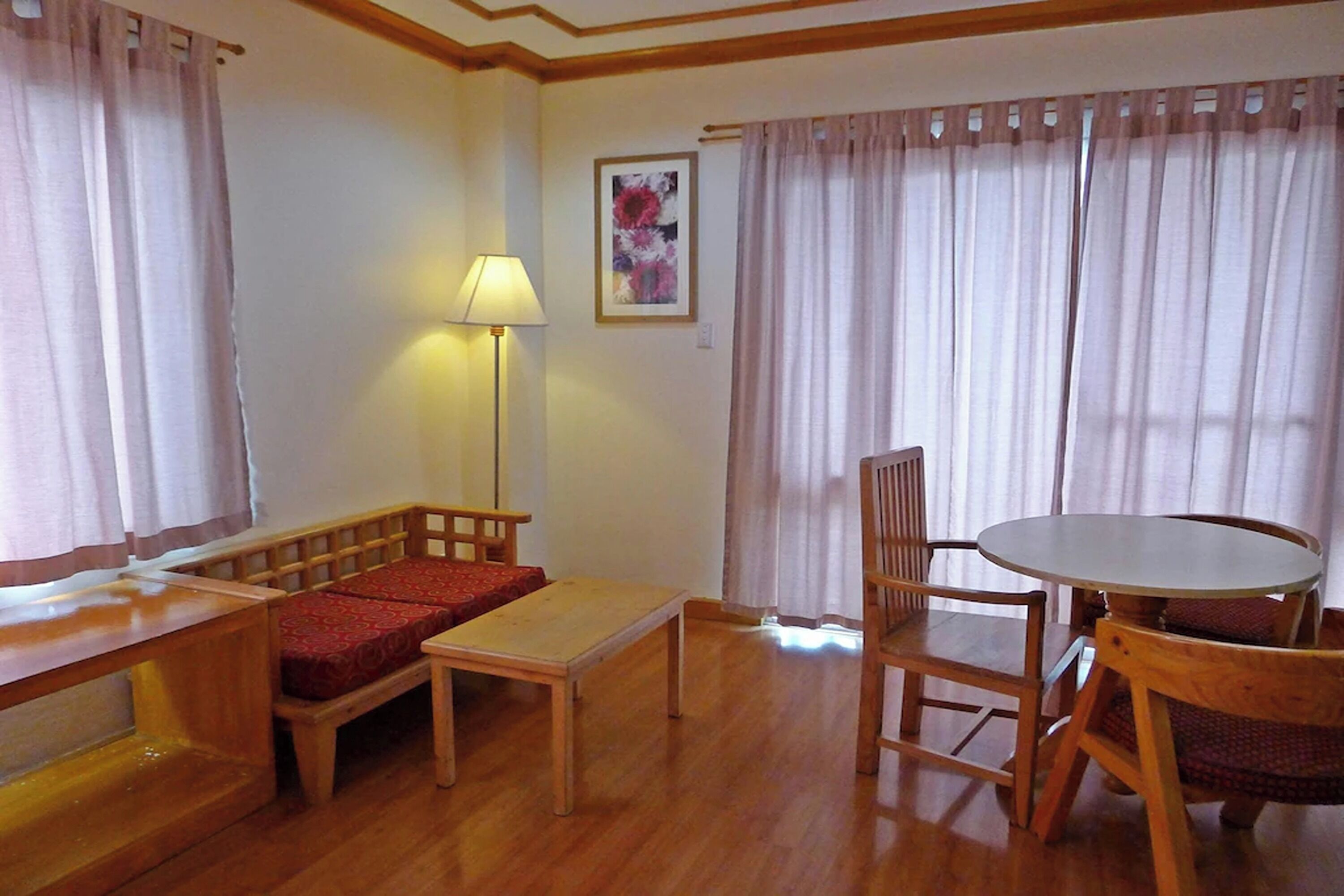 A room with a wooden bench, a small table, a round table with chairs, a lamp, and a painting on the wall.