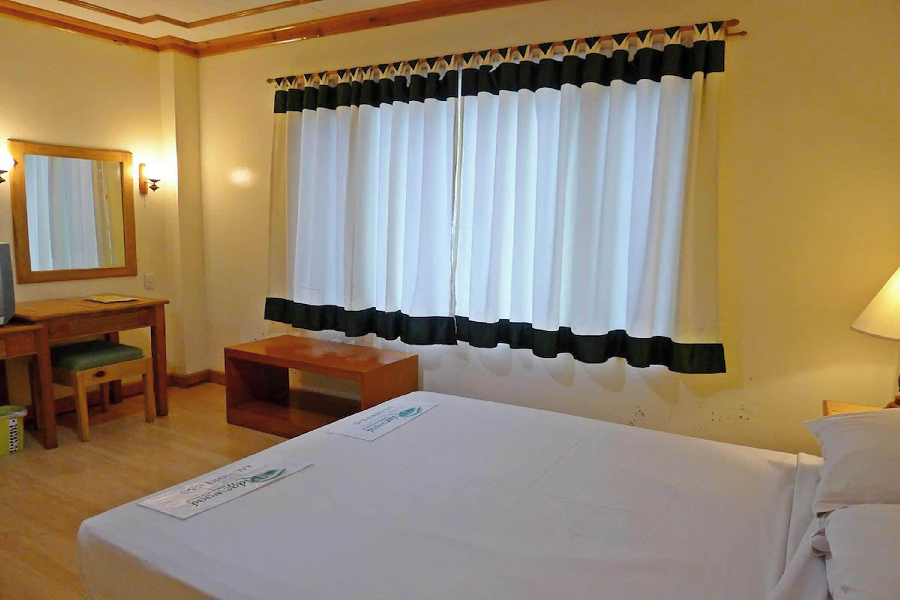 A hotel room with a bed, a desk with a chair, a small table, and a window with curtains.