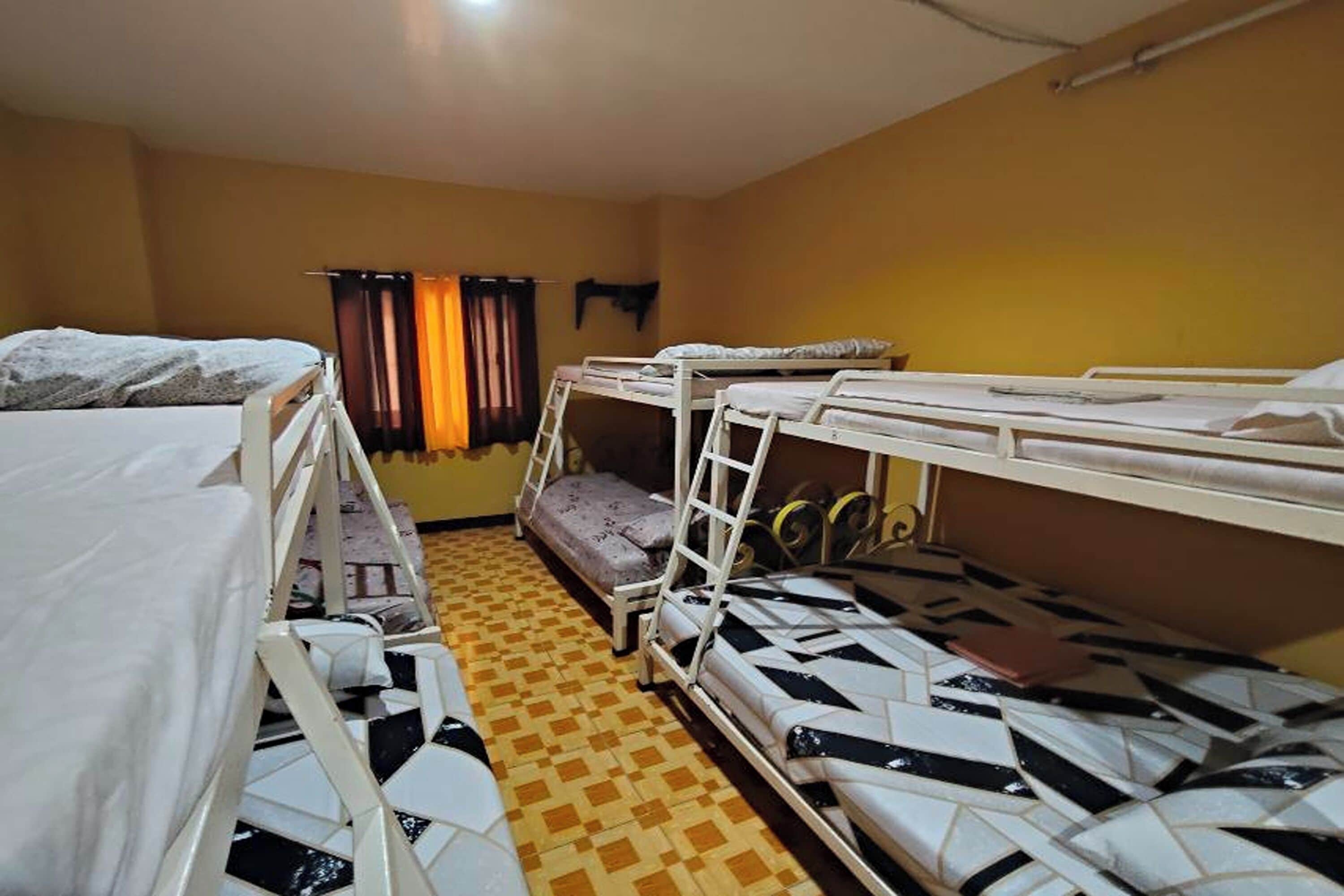 A dormitory room with bunk beds, a patterned floor, and a window with curtains.