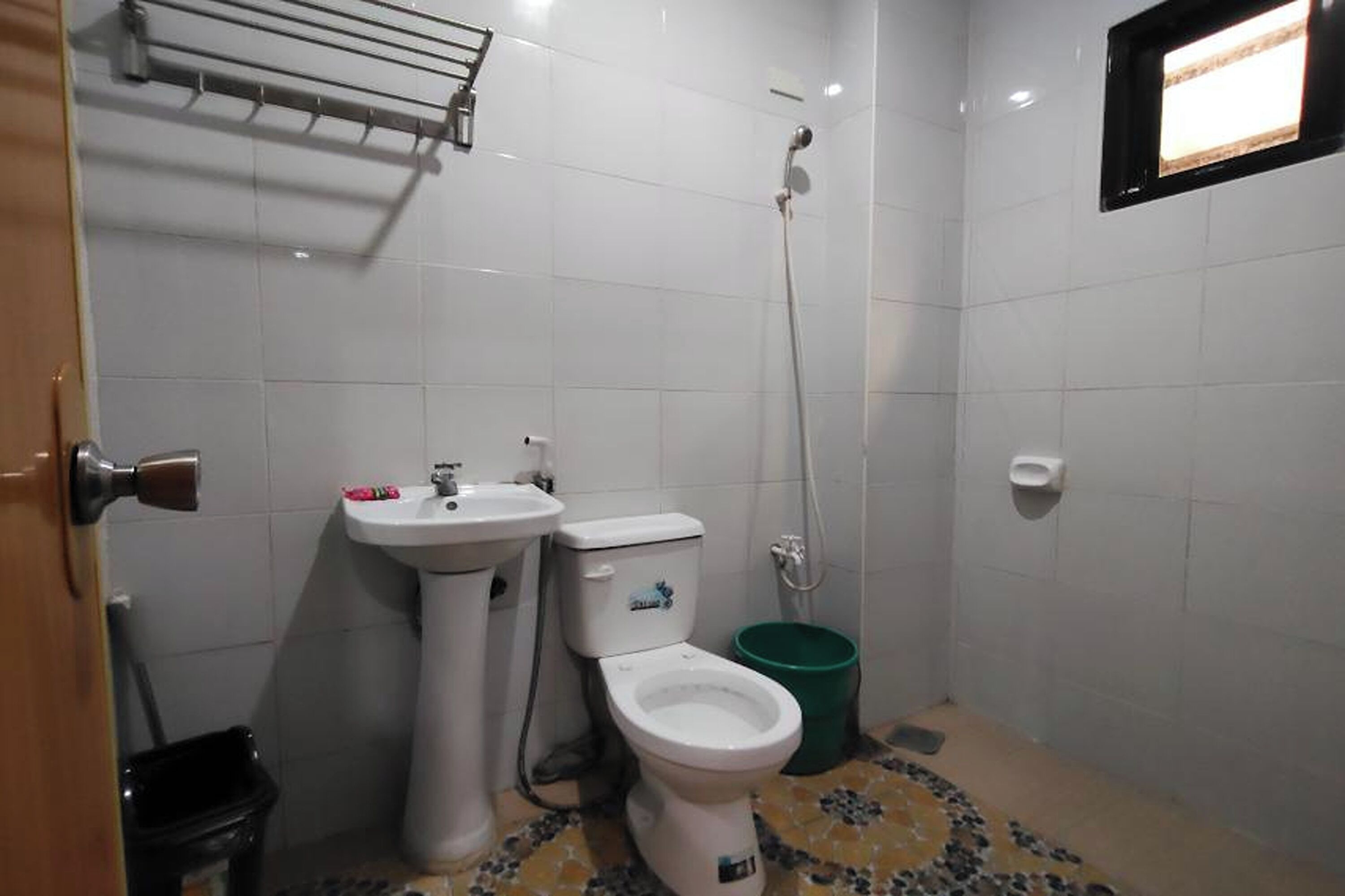 A small bathroom with a toilet, sink, and shower. There is a green trash bin and a towel rack on the wall.