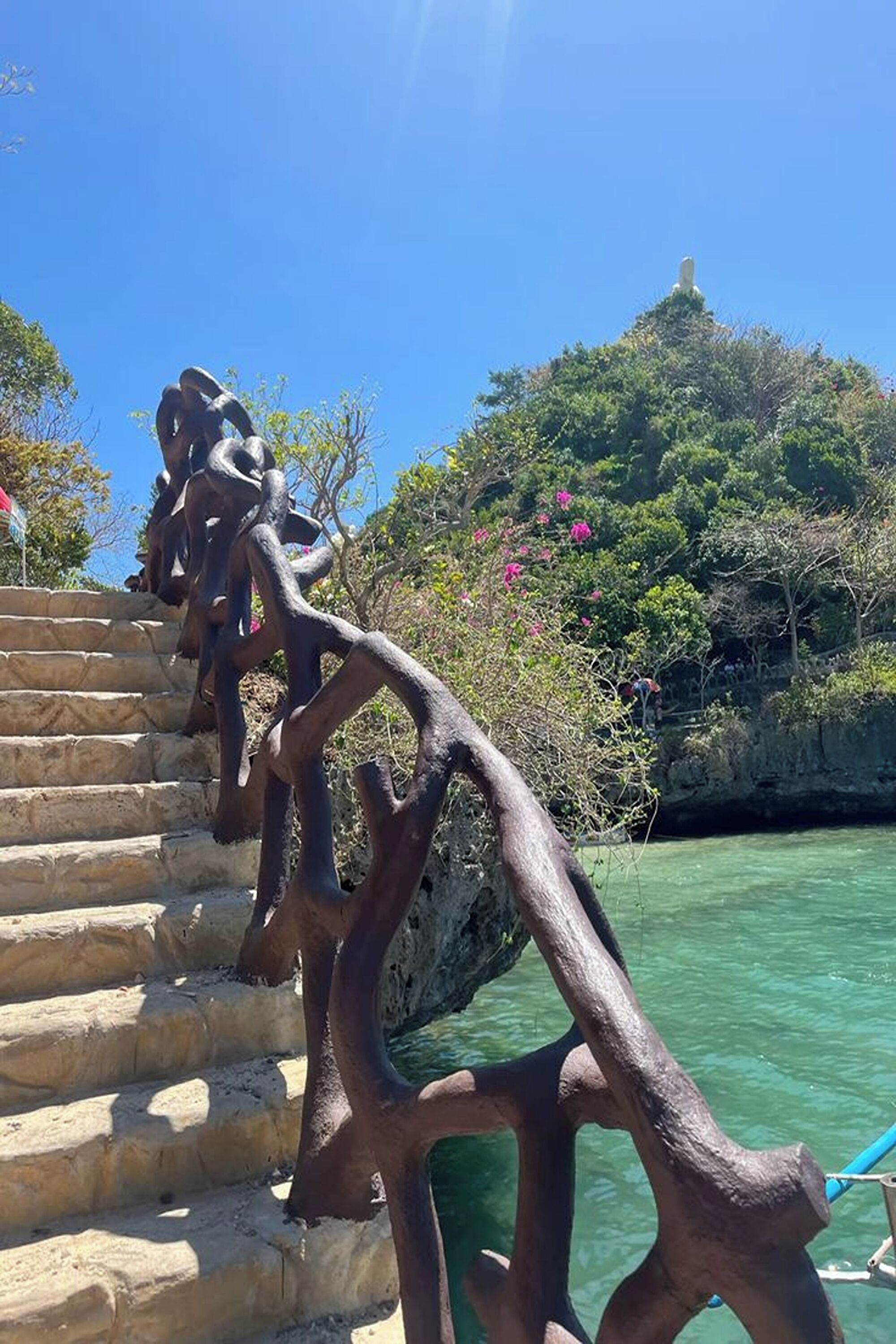 Uma escadaria com grades de metal abstratas e originais, que dá acesso a uma área verde exuberante e com águas cristalinas.