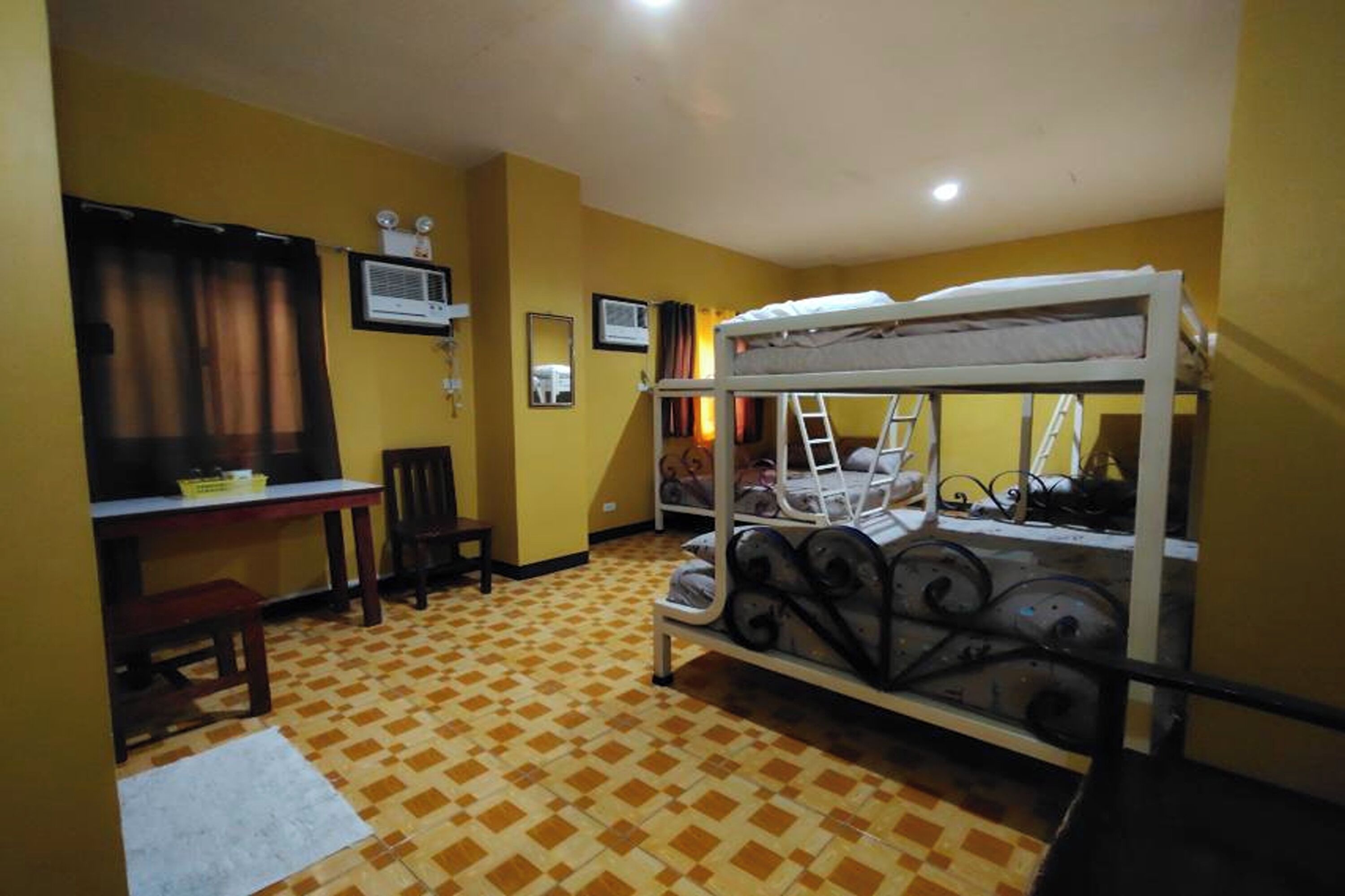 A room with a bunk bed, a desk with a chair, a small table, and a window with curtains.