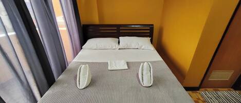A neatly made bed with a grey mattress and white pillows, a wooden headboard, and a window with curtains.
