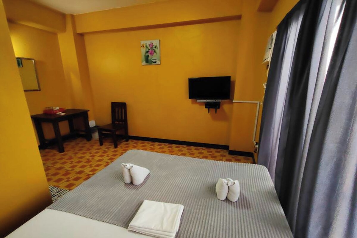Quarto de hotel com cama, televisão na parede, uma cadeira e uma mesinha com um vaso de flores.