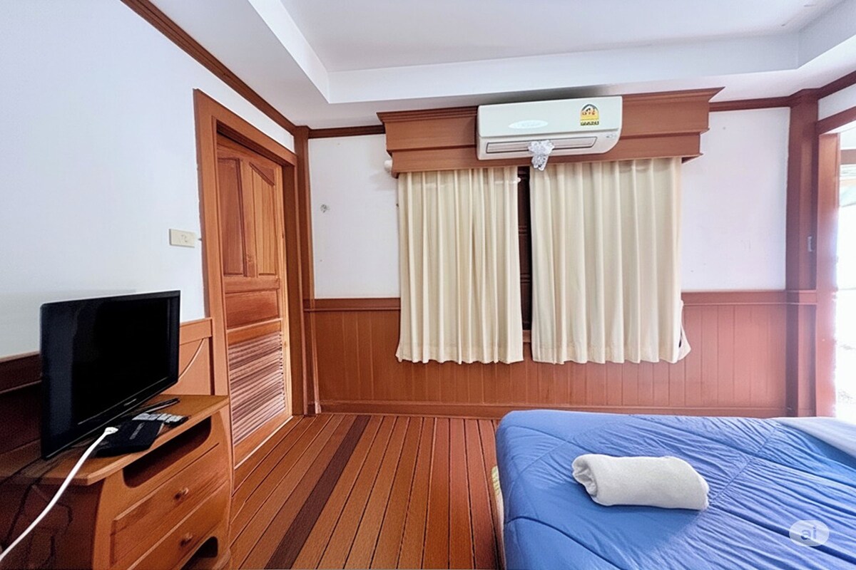 Une chambre d’hôtel avec un plancher en bois, un lit avec un matelas bleu, un téléviseur posé sur un meuble en bois et un appareil de climatisation installé au-dessus de la fenêtre.
