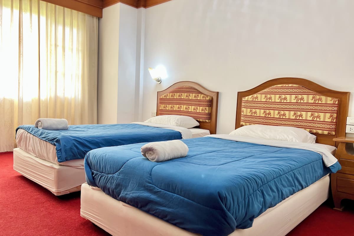 Quarto de hotel com duas camas, cada uma com roupa de cama azul e almofadas brancas. O quarto possui cabeceiras de madeira com estampas de elefantes, um tapete vermelho e uma janela com cortinas.