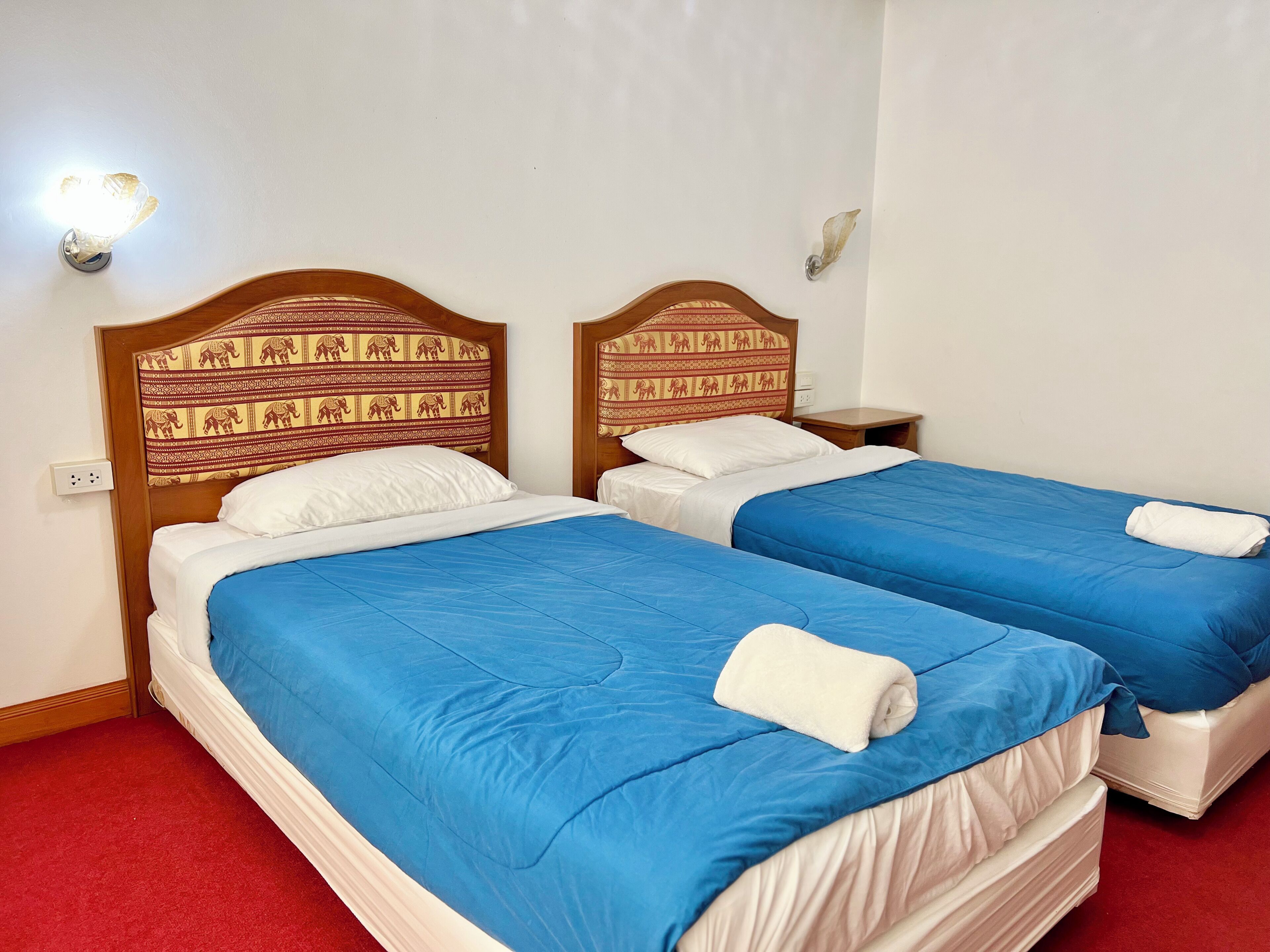 A hotel room with two beds, each with blue bedding and white pillows, wooden headboards with elephant patterns, and a red carpet.