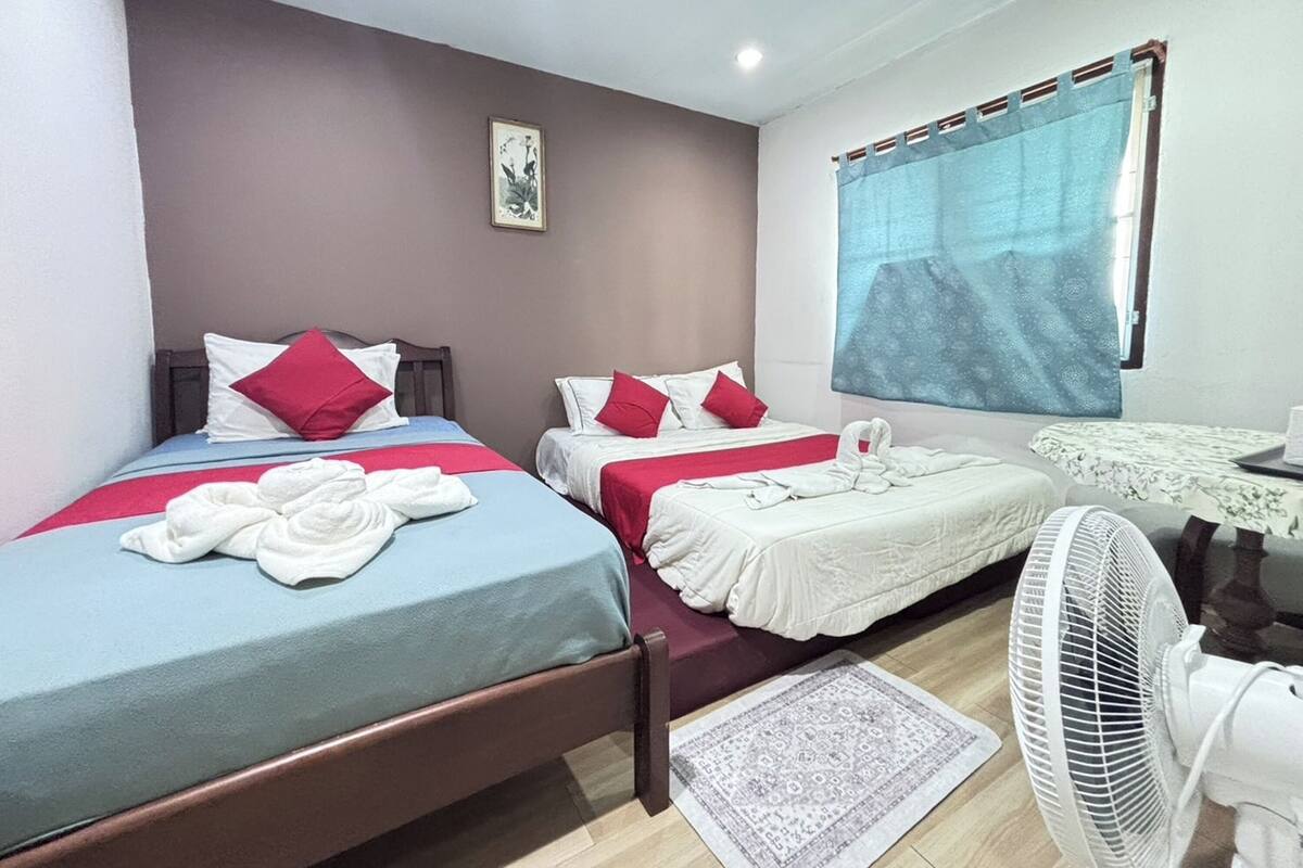 A hotel room with two beds, each with a red and white pillow, a blue bedspread, and a white blanket. There is a small table with a white tablecloth, a fan, and a window with a blue curtain.