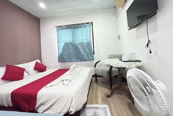 A hotel room with a bed, a small table with a fan, a TV mounted on the wall, and a window with a curtain. - Sunshiny-BB Hostel near Punnavithi BTS station (Bangkok)