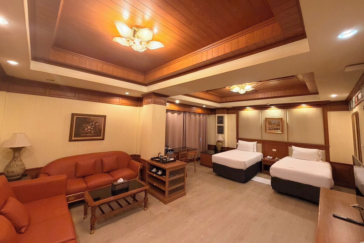 A hotel room with a wooden ceiling, two beds, a sofa, a coffee table, and a lamp.