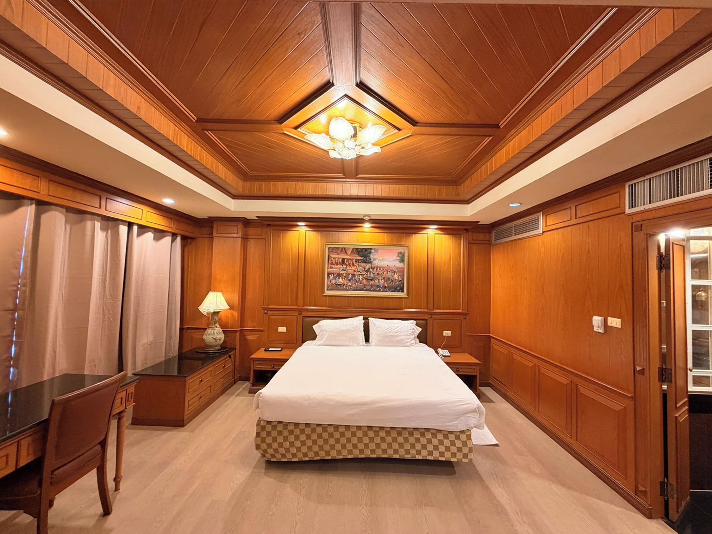 A hotel room with a large bed, wooden paneling, a painting on the wall, and a chandelier.