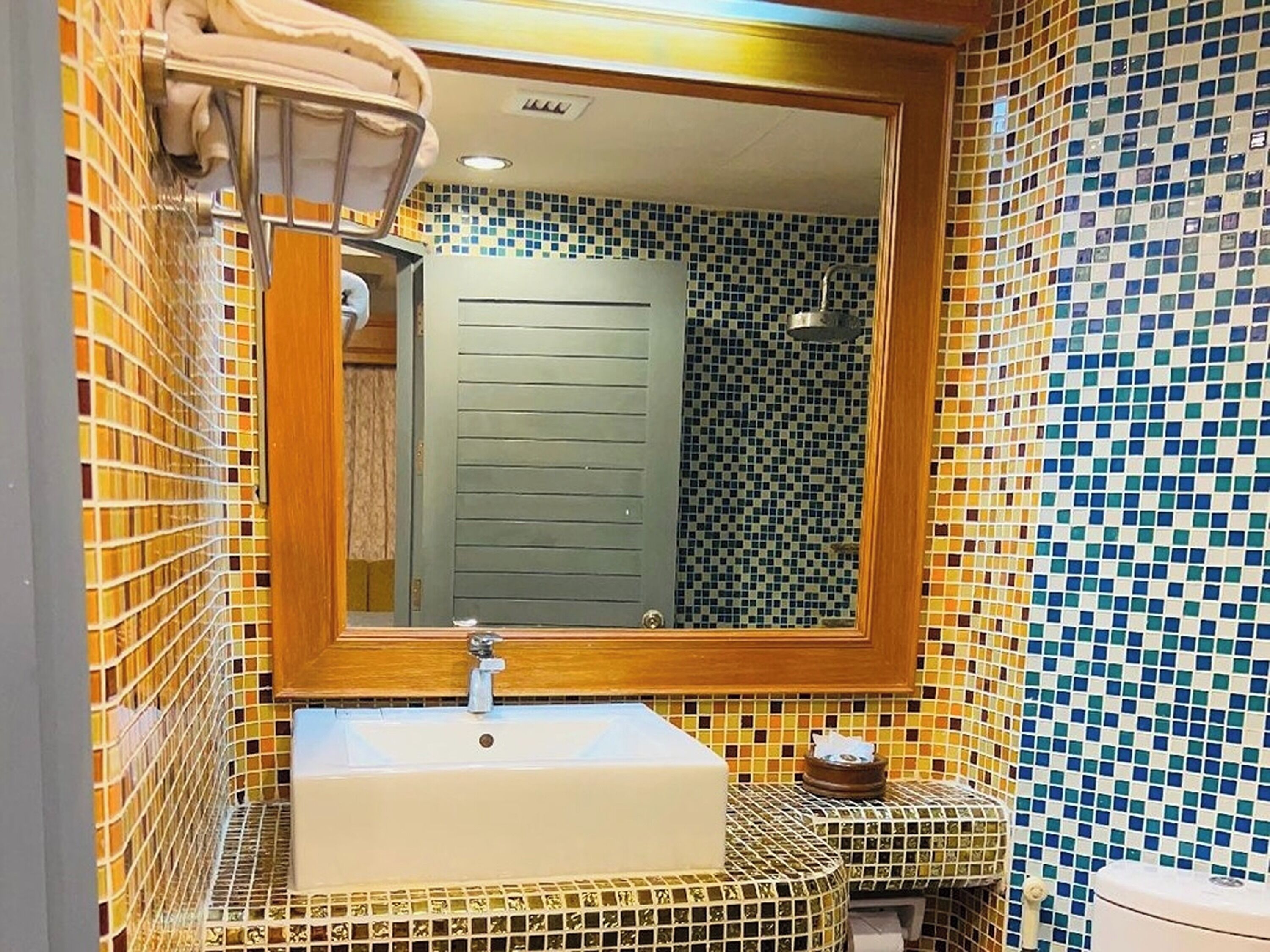 Une salle de bain avec un mur recouvert d’une mosaïque, un miroir à cadre en bois et un lavabo blanc surmonté d’un porte-serviettes.