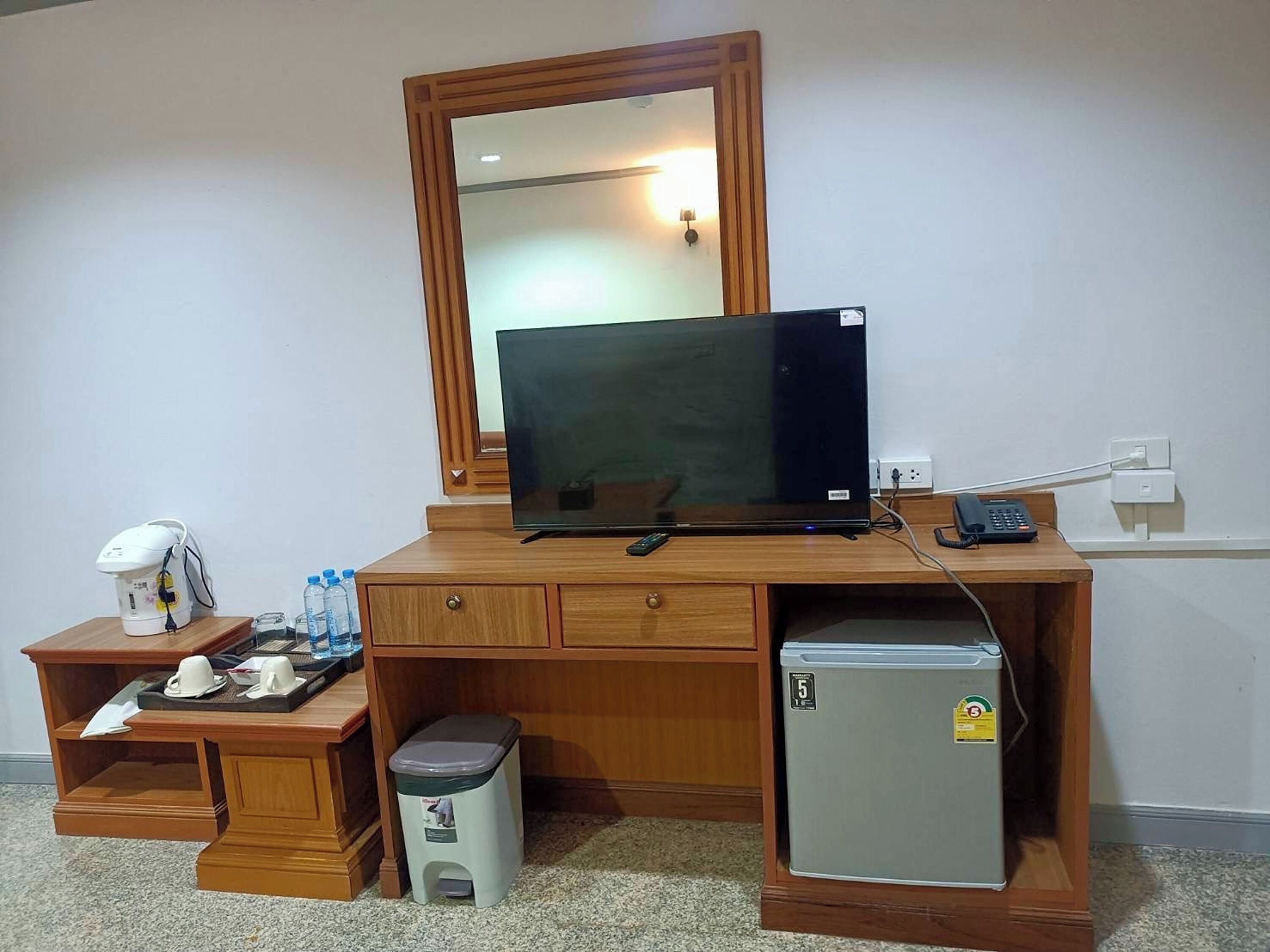 A hotel room with a wooden desk, a television, a mini-fridge, a trash bin, a coffee maker, and a telephone.