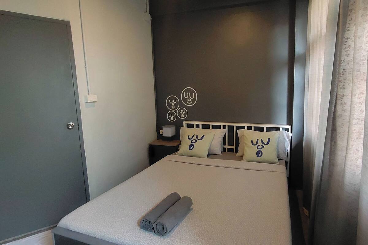 A neatly made bed with a rolled towel, a headboard with decorative wall art, and a curtain on the window.