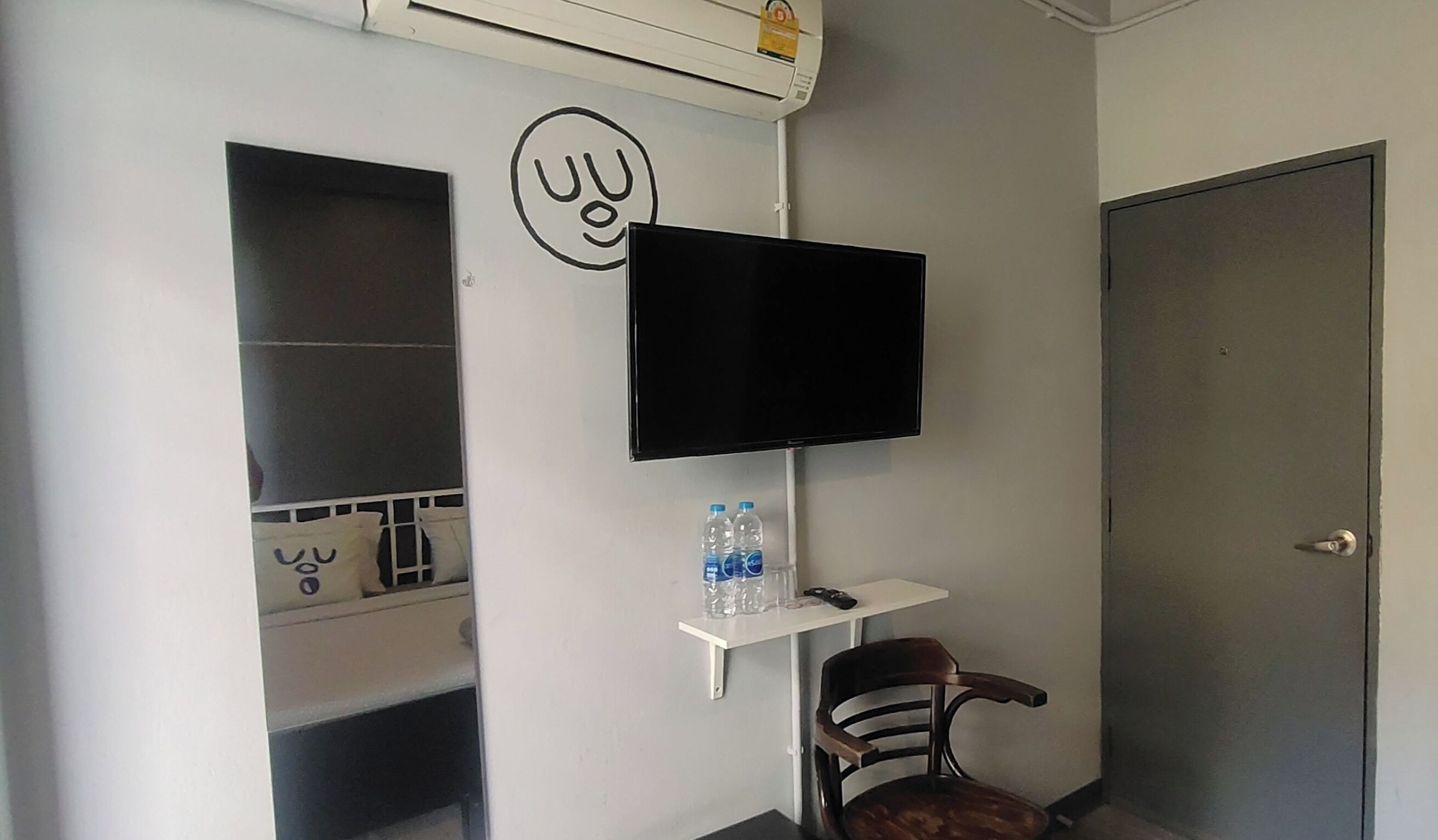 A small room with a mounted TV, a chair, a shelf with water bottles, and a door.