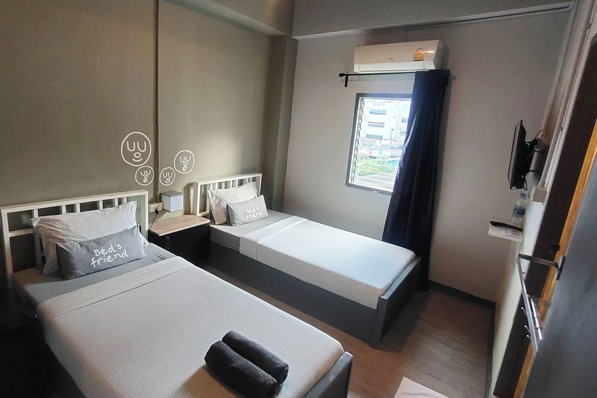 A hotel room with two single beds, a window with curtains, an air conditioning unit, and a bedside table with a lamp.