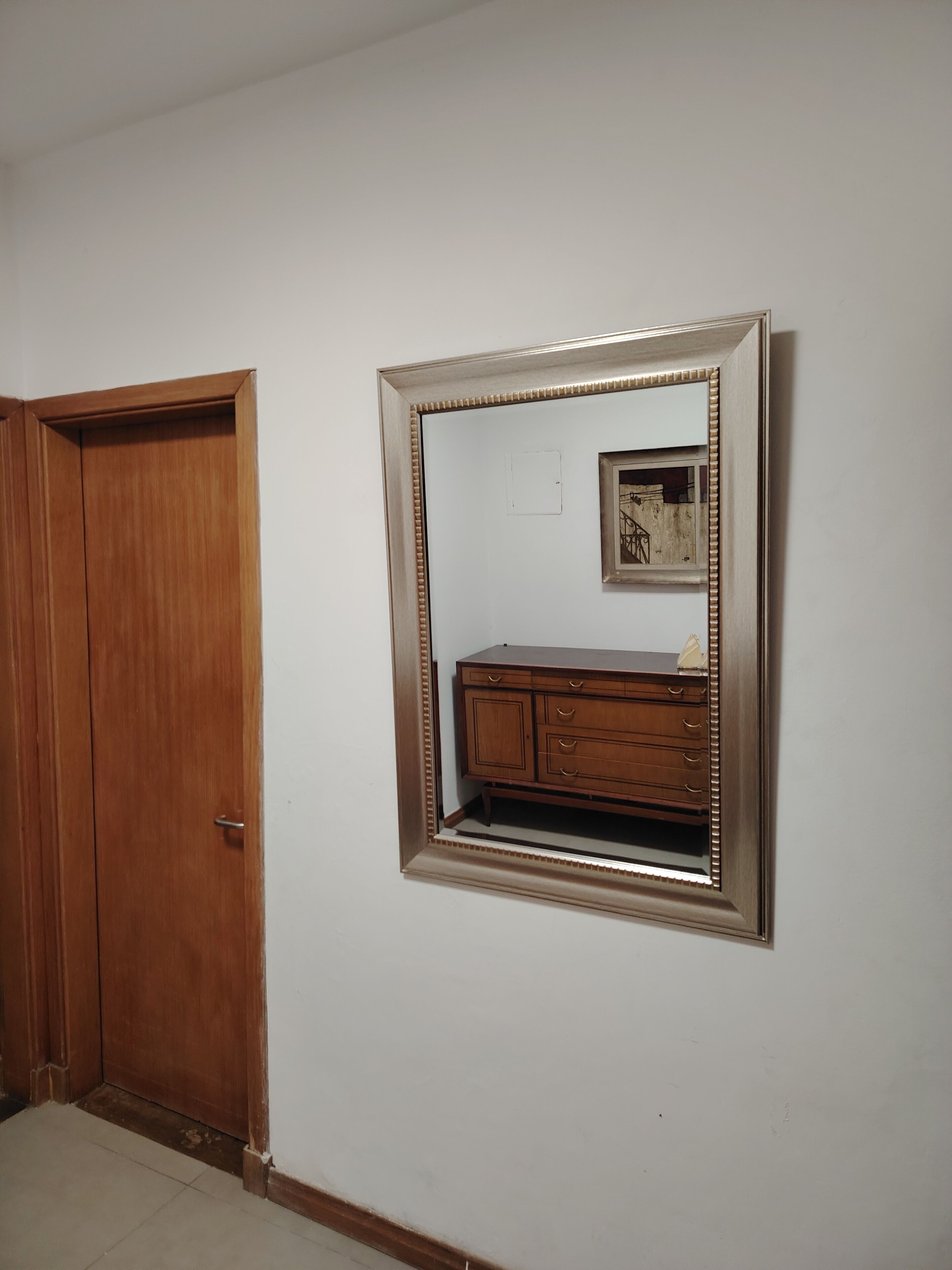 Una habitación con una puerta de madera, un espejo con un marco decorativo y un cuadro pequeño en la pared.