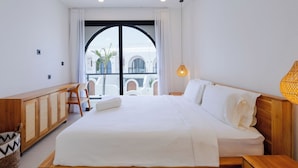 A modern hotel room with a large bed, wooden headboard, and a view of a courtyard with arches. - Villa Amal 9 by Pertama Stay (Canggu)