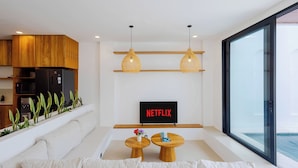A modern living room with a sofa, two wooden tables, a TV displaying 'NETFLIX', and a large window overlooking a body of water. - Villa Amal 9 by Pertama Stay (Canggu)