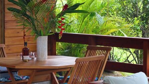 Una terraza de madera con una mesa redonda y sillas, rodeada de exuberante follaje tropical y una pared de madera.