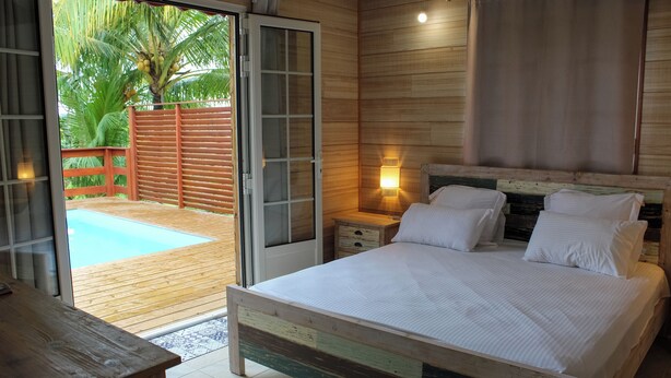 Un dormitorio con cama de madera, mesita de noche, lámpara de pared y puerta corredera de cristal que da a una zona de piscina.